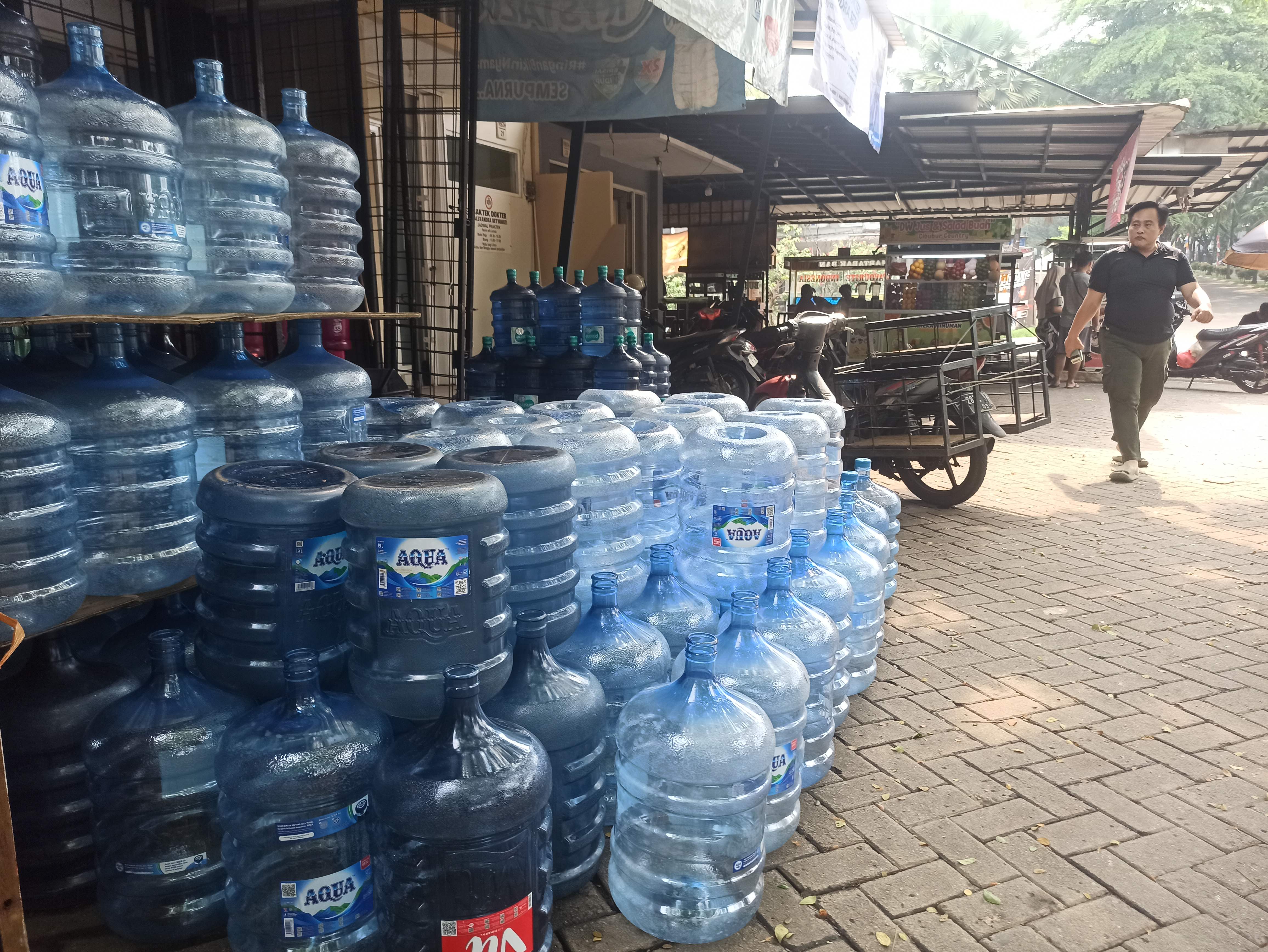 Seorang warga sedang melintas di depan depot isi ulang air mineral yang di depannya berisi tumpukan galon baru dan galon lama berada di kawasan Cibubur, Kota Depok, Jawa Barat. Foto: Apakabar.co.id/Bethriq Kindy Arrazy